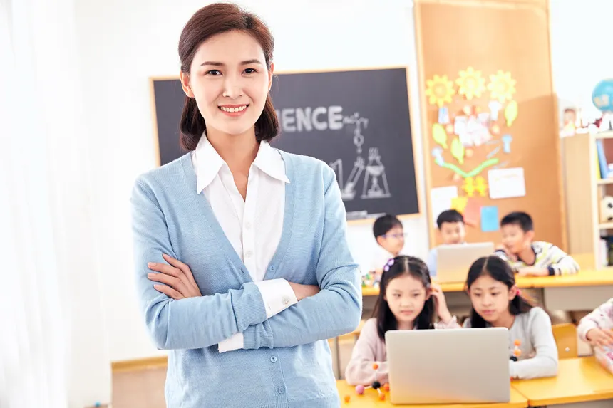 Smiling teacher posing in a classroom with engaged students on laptops.