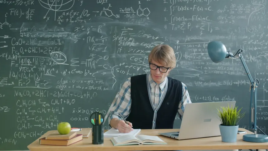 Teacher in classroom writing on notebook, surrounded by books, laptop, and chalkboard.