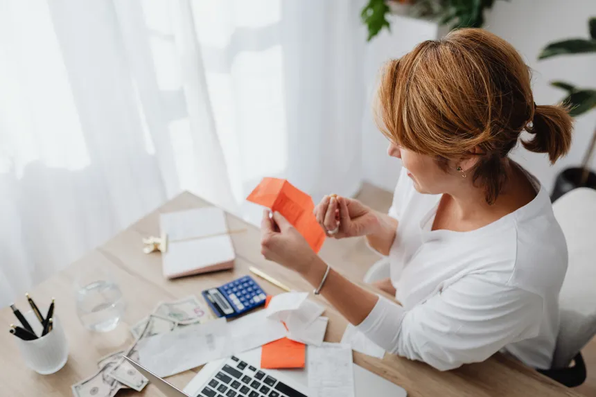A woman organizing her finances with bills and receipts at a home office desk using a calculator and laptop.