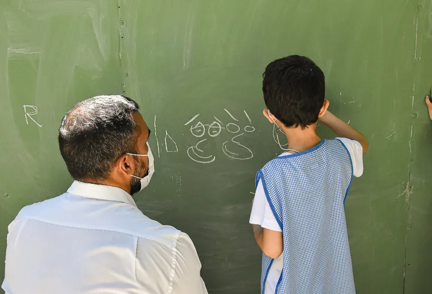 A teacher guides a student writing on a green chalkboard in a classroom setting.