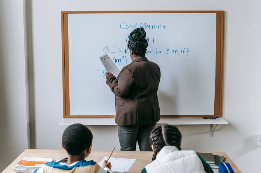 Back view unrecognizable pupils sitting at desk and listening to female teacher writing information on whiteboard