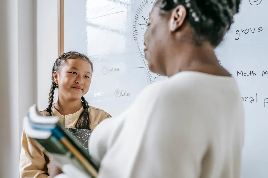 Crop black woman with textbooks standing near whiteboard with ethnic teen girl during lesson in school