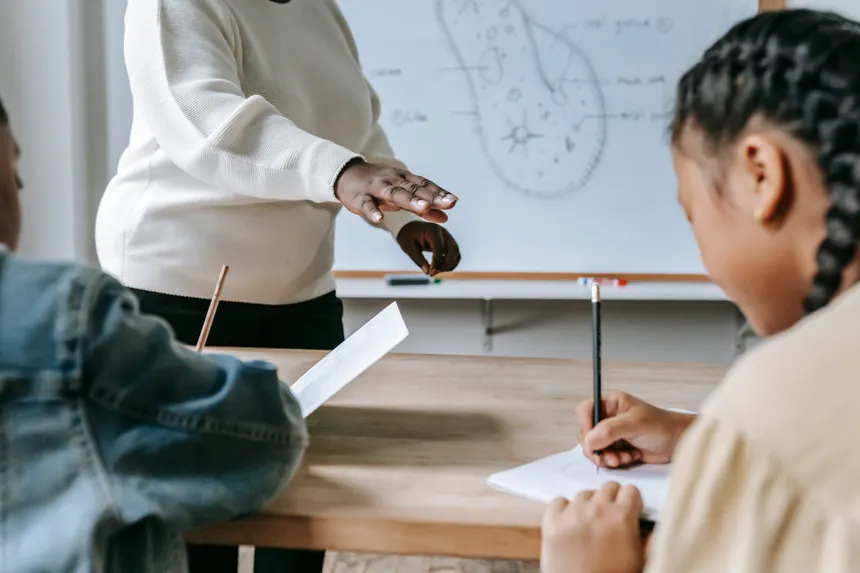 Crop faceless female teacher standing near desk while schoolchildren writing task on papers during lesson