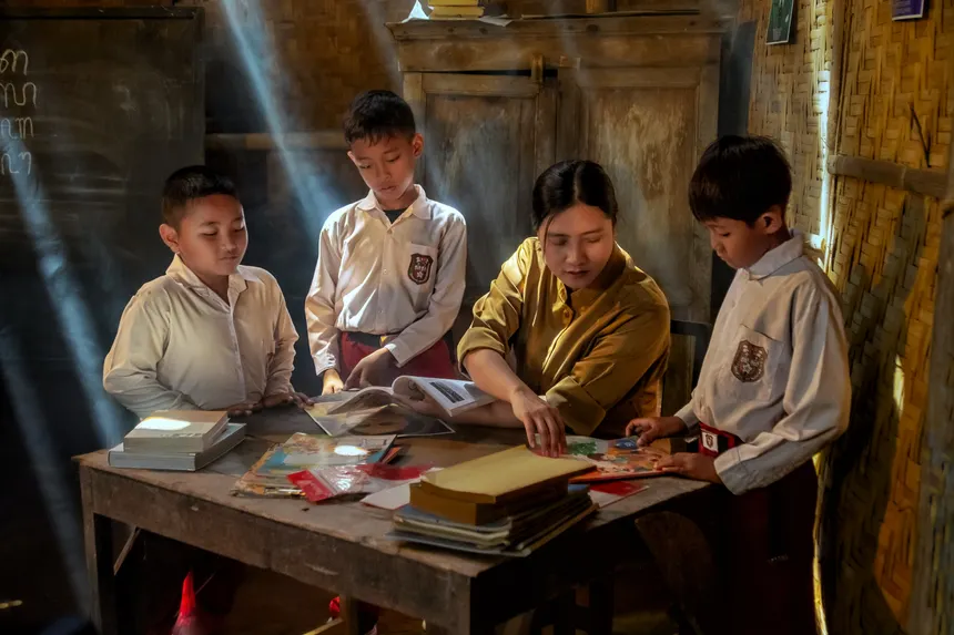 A teacher guiding three students in a rustic classroom, illuminated by sunlight.