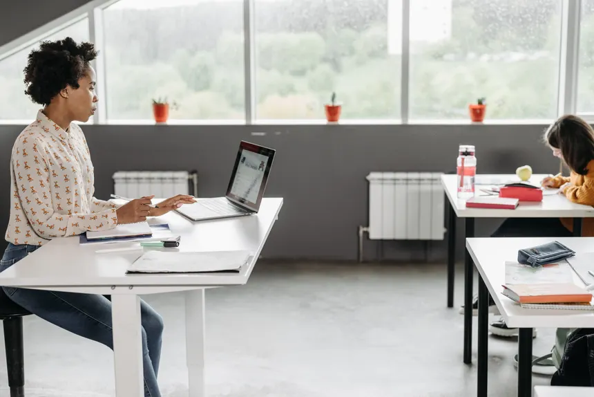 A teacher focused on her laptop in a bright, modern classroom setting.