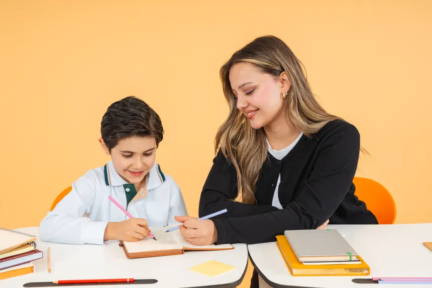 Young teacher helps a schoolboy with writing at a desk, fostering learning.